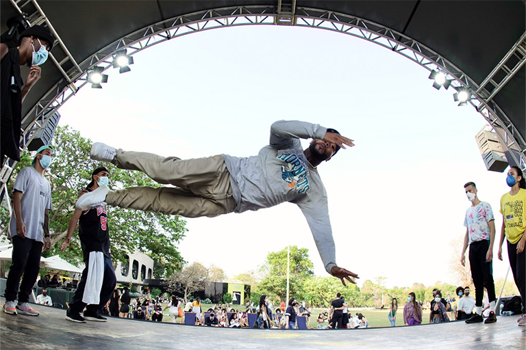 Dançarino realiza um movimento aéreo de breakdance em um palco ao ar livre, observado por outros participantes e público ao fundo.