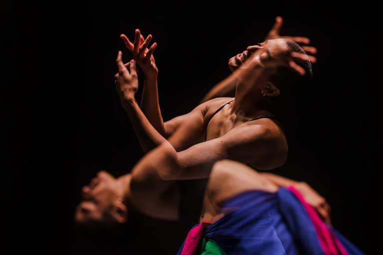 Pessoa dançando com expressão intensa e braços em movimento sob iluminação dramática.