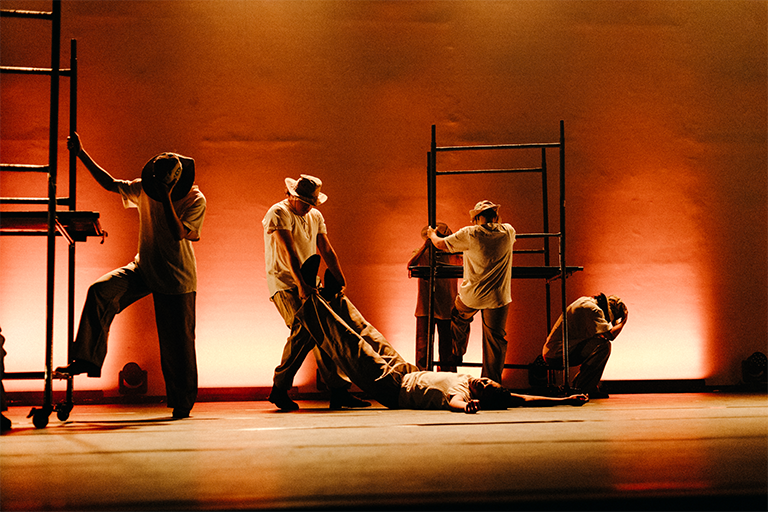 Grupo de dançarinos em cena teatral, com iluminação âmbar e estruturas metálicas no palco, representando esforço e movimento coletivo.