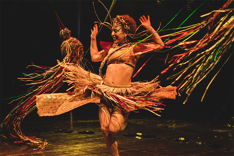 Pessoa dançando com expressão alegre, vestida com traje artesanal de palha e fitas coloridas, em palco iluminado.